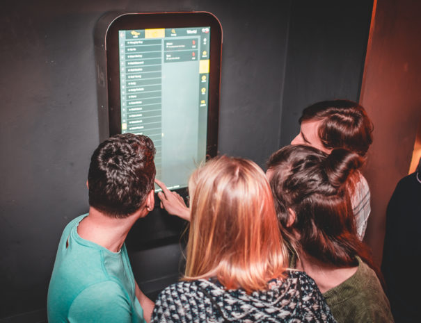 Four people together around a touchscreen karaoke jukebox in the full stream of a party.